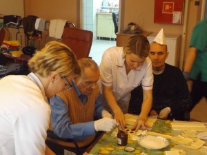 Uśmiechnięci pensjonatiusze wraz z personelem w czasie wypiekania smacznych ciasteczek w czapeczkach z papieru